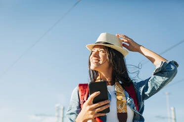 Happy woman wearing hat and standing with smart phone under sky - DCRF01730