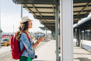 Junge Frau mit Hut, die an einer Straßenbahnhaltestelle einen Streckenplan durchgeht - DCRF01734