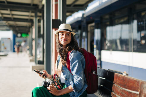 Glückliche Frau mit Hut sitzt auf einer Bank an einer Straßenbahnhaltestelle - DCRF01736