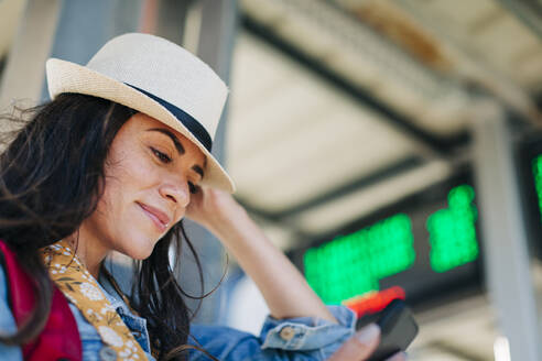 Smiling woman wearing hat using mobile phone at tram station - DCRF01740