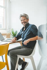 Smiling businessman sitting on chair at workplace - JOSEF20489