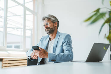 Smiling businessman holding smart phone by laptop at desk in office - JOSEF20496