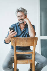 Lächelnder Geschäftsmann hält Smartphone und sitzt auf einem Stuhl im Büro - JOSEF20535