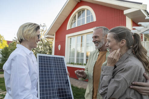 Ein glückliches Paar diskutiert mit einem Agenten, der ein Solarpanel vor seinem Haus hält - VPIF08433