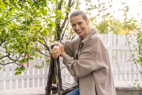 Happy woman leaning on ladder in back yard - VPIF08462