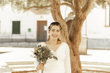 Pregnant bride holding flower bouquet in front of tree - PCLF00651