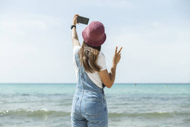 Eine stilvolle Frau hält einen sonnigen Strandmoment mit einem Selfie auf Mallorca, Spanien, fest, wobei sie eine Brille und einen Hut trägt - PCLF00667