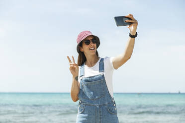 Eine stilvolle Frau macht ein Selfie am sonnigen Strand von Mallorca, Spanien, mit einer Brille und einem trendigen Hut - PCLF00668