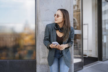 Smiling woman with smart phone standing in front of building - NDEF00962