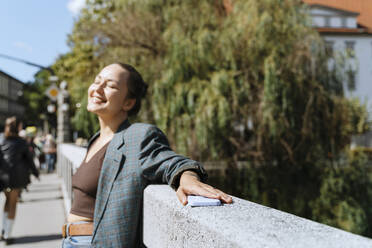Lächelnde Frau verbringt ihre Freizeit an einer Mauer an einem sonnigen Tag - NDEF00974