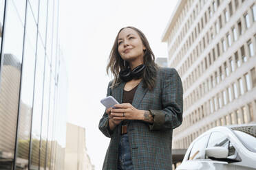 Contemplative woman standing with smart phone near building - NDEF00980