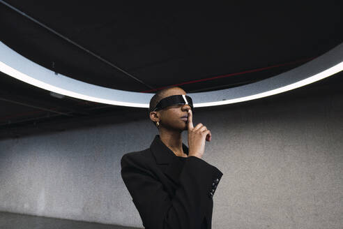 Young woman with finger on lips using futuristic glasses in parking garage - PNAF05961