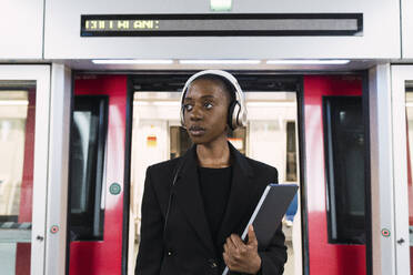 Nachdenkliche Frau mit Kopfhörern und einem Laptop vor der U-Bahn stehend - PNAF06004