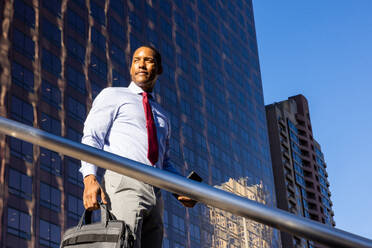 Gutaussehender afrikanischer Geschäftsmann, CEO, in einem eleganten Anzug in einem Geschäftszentrum im Freien - Schwarzer männlicher Pendler auf dem Weg zur Arbeit, Stadt und Finanzviertel im Hintergrund - DMDF01563
