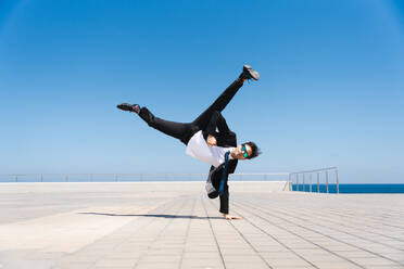 Happy and handsome adult businessman wearing elegant suit doing acrobatic trick moves in the city, alternative concept for business advertisement with energetic and creative people - DMDF01578