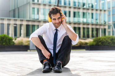 Happy and handsome adult businessman wearing elegant suit sitting on the ground and listening music with headphones to relax - DMDF01610