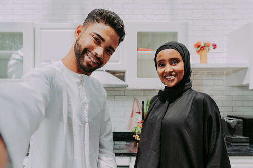 Happy middle-eastern couple wearing traditional arab clothing at home - Married arabian husband and wife bonding together in the apartment, concepts about relationship, domestic life and emirati lifestyle - DMDF01623