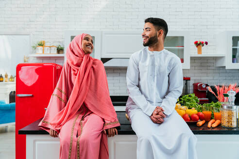 Happy middle-eastern couple wearing traditional arab clothing at home - Married arabian husband and wife bonding together in the apartment, concepts about relationship, domestic life and emirati lifestyle - DMDF01636