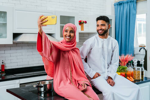 Happy middle-eastern couple wearing traditional arab clothing at home - Married arabian husband and wife bonding together in the apartment, concepts about relationship, domestic life and emirati lifestyle - DMDF01637