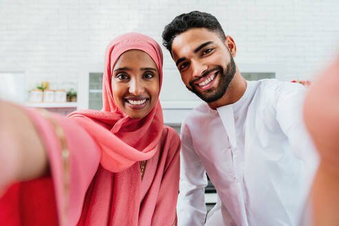 Happy middle-eastern couple wearing traditional arab clothing at home - Married arabian husband and wife bonding together in the apartment, concepts about relationship, domestic life and emirati lifestyle - DMDF01639