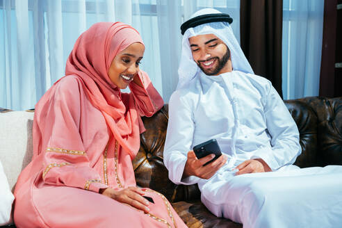 Happy middle-eastern couple wearing traditional arab clothing at home - Married arabian husband and wife bonding together in the apartment, concepts about relationship, domestic life and emirati lifestyle - DMDF01641