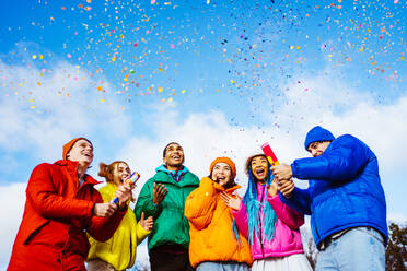 Multiracial group of young happy friends meeting outdoors in winter and celebrating party with confetti shooter, wearing winter jackets and having fun - Multiethnic millennials bonding in a urban area, concepts about youth and social releationships - DMDF01952