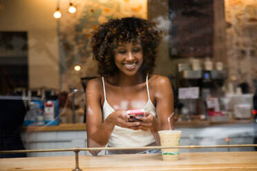 Schöne Afro-Frau, die in einem Kaffeehaus sitzt - Hübsches Mädchen mittleren Alters, das in einer Bar Kaffee trinkt, Reflexionseffekt vom Fensterglas - DMDF02054