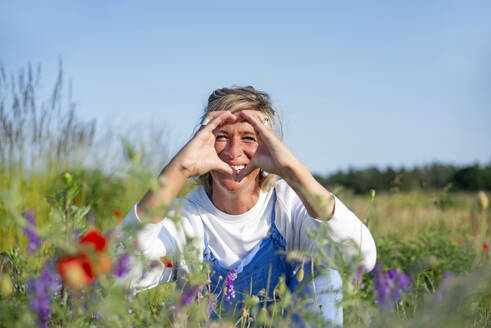 Happy woman making heart gesture sitting in meadow - BFRF02433