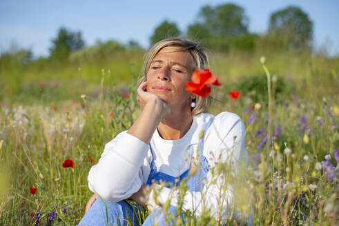 Frau mit Hand am Kinn sitzt inmitten von Blumen auf einer Wiese - BFRF02438