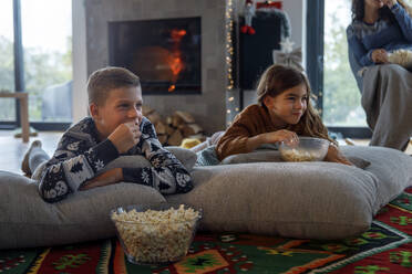 Lächelnde Geschwister, die zu Hause mit ihrer Mutter fernsehen und Popcorn essen - IKF01089