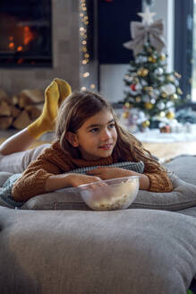 Girl having popcorn lying on pillows in living room - IKF01092