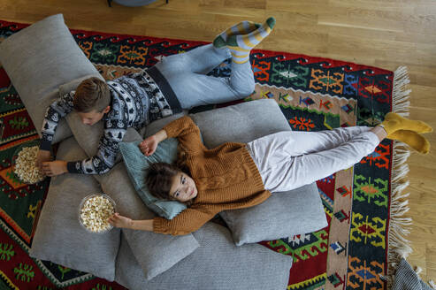 Siblings with popcorn bowls lying on pillows at home - IKF01104