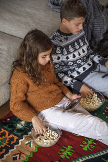 Siblings using smart phones and eating popcorn at home - IKF01118