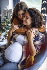 Girl embracing expectant mother sitting on chair at home - IKF01130