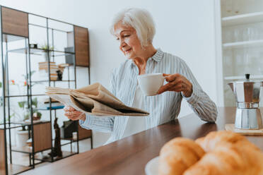 Senior woman relaxing at home, reading newspaper and having breakfast - DMDF02454