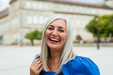 Porträt einer positiven, modernen älteren Frau im blauen Kleid, die auf einer Straße in der Stadt steht und fröhlich in die Kamera schaut - ADSF46565