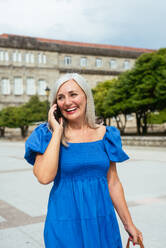 Smiling modern senior female in blue dress speaking on mobile phone on city street against blurred background - ADSF46568