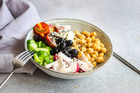 High angle of bowl with healthy salad and vegetables with chickpea sesame seeds and olives with cutlery and fabric placed on bright gray table - ADSF46573