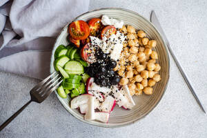 Top view of bowl with healthy salad and vegetables with chickpea sesame seeds and olives with cutlery and fabric placed on bright gray table - ADSF46574