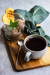 High angle of glass of homemade coffee ice cream with waffle and fresh mint near spoon and cup of coffee placed on wooden tray with napkin against gray surface - ADSF46577