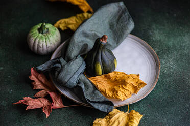 High angle of autumnal table setting with napkin, leaves and pumpkins placed on plate against dark surface - ADSF46580
