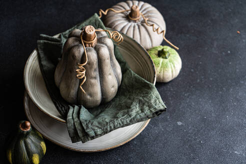 High angle of autumnal table setting with napkin and pale colored pumpkin placed on ceramic bowl near pale green pumpkins against dark surface - ADSF46582
