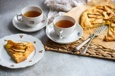 High angle of plates with pieces of apple pie and cups of tea near knife, fork and napkin on rustic tray against gray table - ADSF46587