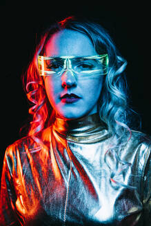 Portrait of cybernetic young girl with curly hair and futuristic glasses wearing bright and reflecting clothing looking at camera while standing against black background - ADSF46591