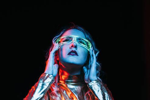 From below of cybernetic young girl with curly hair wearing bright and reflecting clothing looking away while standing and adjusting futuristic glasses against black background - ADSF46592