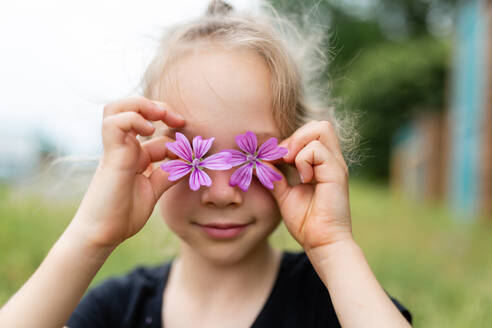 Portrait of cute little girl covering eyes with purple delicate mallow flowers in summer time - ADSF46593