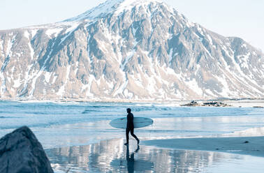 Side view of unrecognizable male surfer in wetsuit carrying surfboard while walking on sandy beach near reflective wavy sea with snowy mountain in Norway - ADSF46597