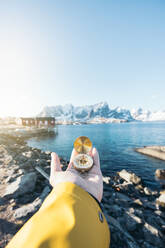 Anonymous traveler in warm yellow jacket while standing with compass in hand on rocky edge of sea against buildings snowy mountains under cloudless blue sky in Lapland in sunny daylight - ADSF46599