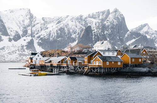 Typical small fishermen village with yellow wooden cabin houses built on seaside of fjord seawater in Lapland against snowy mountains under cloudless blue sky in daylight - ADSF46600