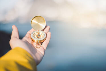 Anonymous traveler in warm yellow jacket while standing with compass in hand on rocky edge of sea against blurred background in Lapland in sunny daylight - ADSF46602